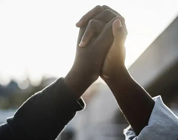 Two people raising and clasping hands together outdoors, silhouetted against bright sunlight.